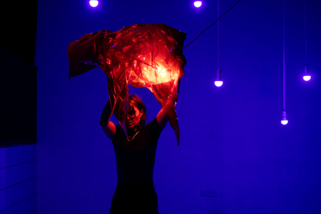 The photo shows performer Rita Mazza in a stage or rehearsal setting illuminated with intense, colored lighting. The background glows in deep blue-purple, with several hanging light bulbs emitting a vivid violet light. Rita Mazza stands in the foreground, mostly in shadow, holding a piece of shiny, translucent foil in warm red-orange above her head. A light source behind the foil makes it glow brightly, creating a strong contrast against the cool tones of the background. The scene feels theatrical, mysterious, and full of energy.