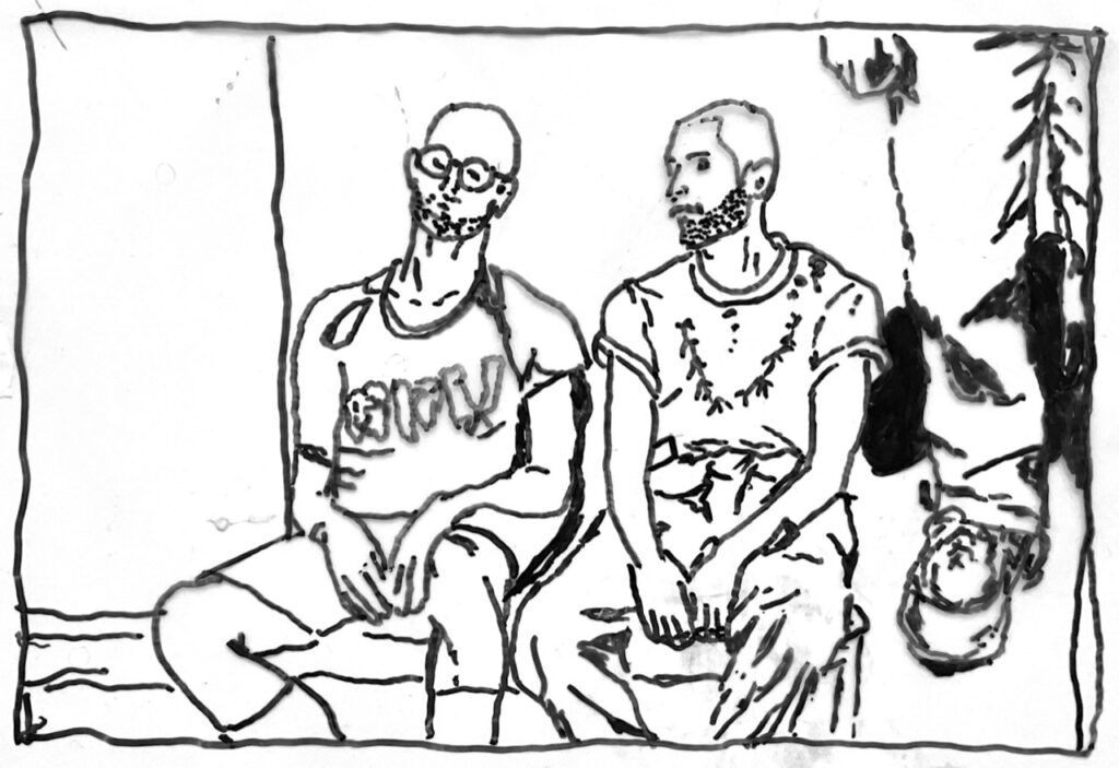 Das Bild zeigt eine schwarz-weiße Zeichnung von drei Personen.
Zwei Personen sitzen nebeneinander auf einer Bank oder einem Bett. Sie haben kurze Haare und Bärte. Die linke Person trägt eine Brille und ein T-Shirt mit einem Schriftzug, die rechte Person ein T-Shirt mit Muster. Beide sitzen mit den Händen auf den Oberschenkeln.
Rechts im Bild hängt eine dritte Person kopfüber, ebenfalls mit kurzem Haar und Bart.
Der Zeichenstil ist sehr reduziert, mit einfachen, dünnen Linien und wenig Details.