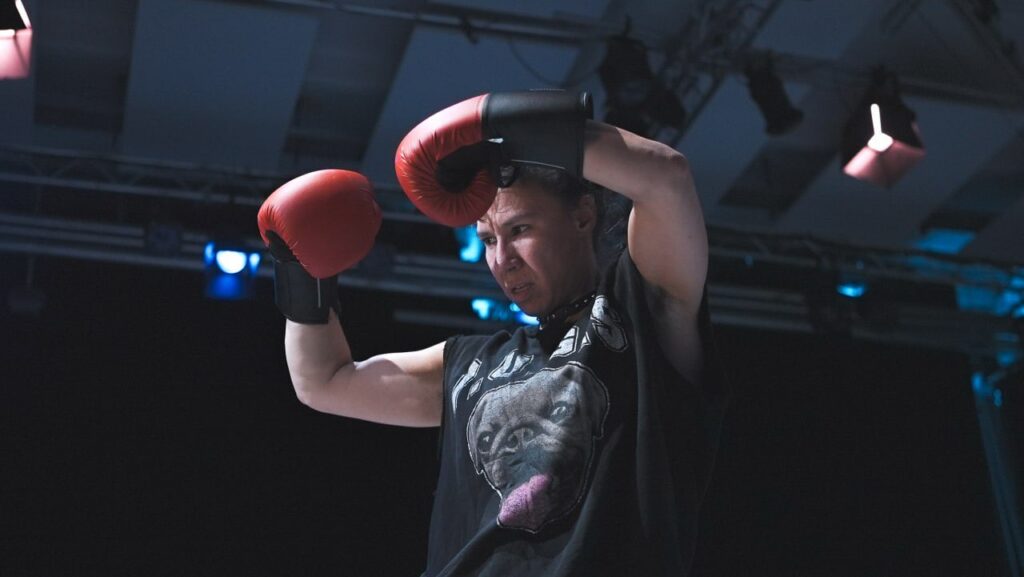 Das Bild zeigt Birte Opitz, die ihre mit zwei Boxhandschuhen bestückten Hände gegeneinander presst. Ihr schwarzes Shirt zeigt eine Bulldogge. Im Hintergrund ist die Decke eines Performanceraums zu sehen (mit Lichtern und Lichttrasse).