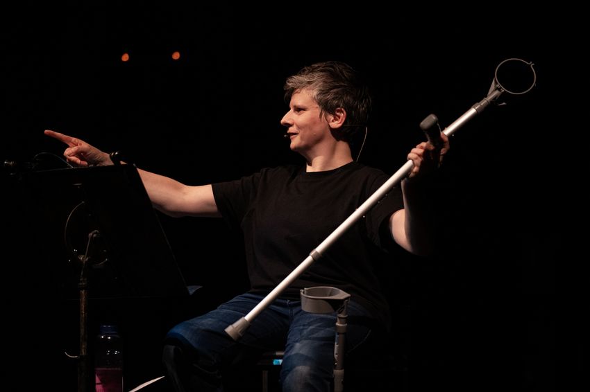 Auf dem Bild ist die Choreografin Claire Cunningham auf einer Bühne zu sehen. Sie trägt ein schwarzes T-Shirt und Jeans und sitzt, leicht seitlich gedreht, auf einem Stuhl. In der rechten Hand hält sie eine Unterarmgehstütze, mit der linken Hand zeigt sie nach vorne. Vor ihr steht ein Noten- oder Lesepult. Die Szene ist dunkel ausgeleuchtet, sodass sie im Zentrum des Lichts steht, während der Hintergrund schwarz bleibt.