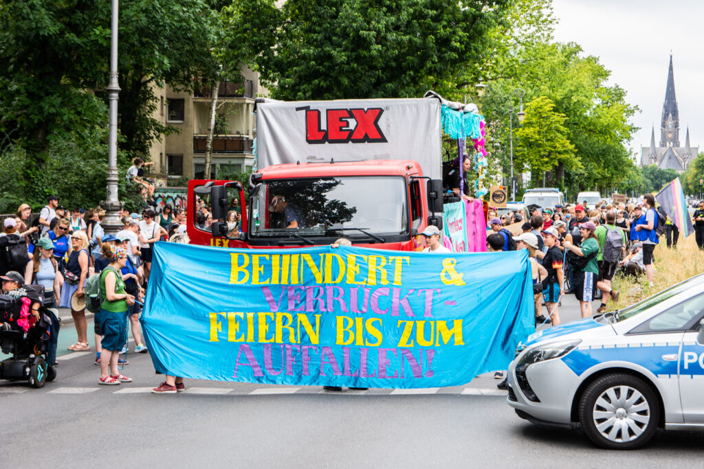 The photo was taken at the behindert und verrückt Pride Parade. It shows a demonstration march led by a truck with a banner stretched across the front. The banner reads, in yellow and pink block letters: “behindert & verrückt - feiern bis zum Auffallen” (Disabled & crazy – celebrate until you stand out!). Behind it is a crowd of people wearing high-visibility vests and waving flags, among other things. A police car can be seen on the right-hand side of the picture.