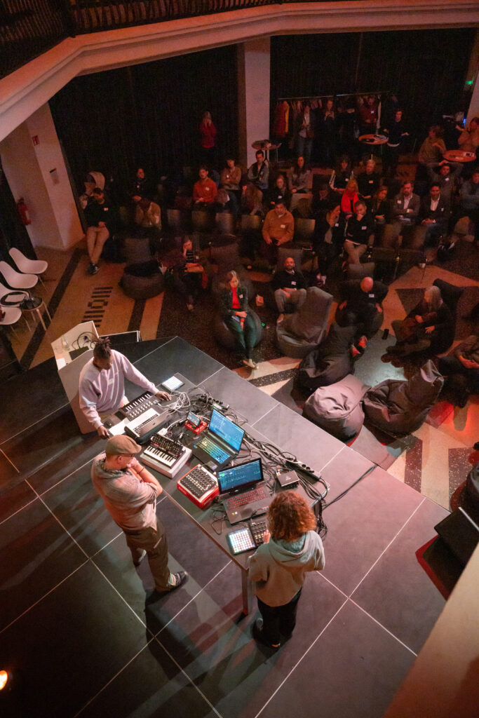 Das Foto zeigt einen Raum mit vielen Menschen. Im Vordergrund stehen drei Personen (sub_bar) an einem Tisch mit mehreren Laptops, Kabeln und Musikgeräten. Sie scheinen elektronische Musik zu spielen oder etwas zu präsentieren. Im Hintergrund sitzen und liegen die Zuschauerinnen und Zuschauer entspannt auf Stühlen und Sitzsäcken. Der Raum ist warm beleuchtet und wirkt gemütlich.